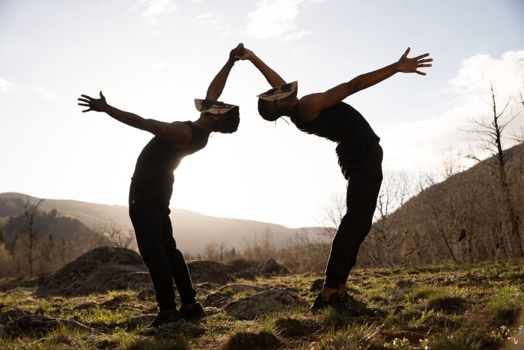 Two men wearing masks bend over backwards towards each other, two of their hands meeting. They're outside and silhouetted against a rising sun.