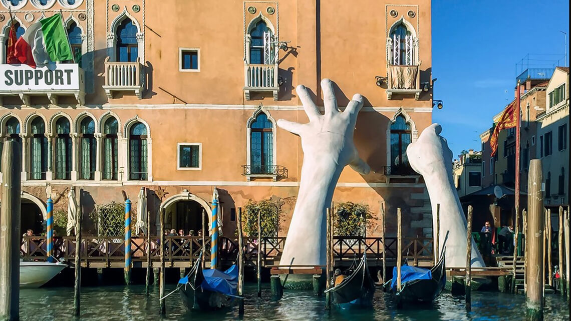 A monumental sculpture of two hands emerging out of a Venetian canal appearing to hold up a building.
