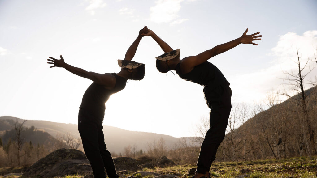 Two men wearing masks bend over backwards towards each other, two of their hands meeting. They're outside and silhouetted against a rising sun.