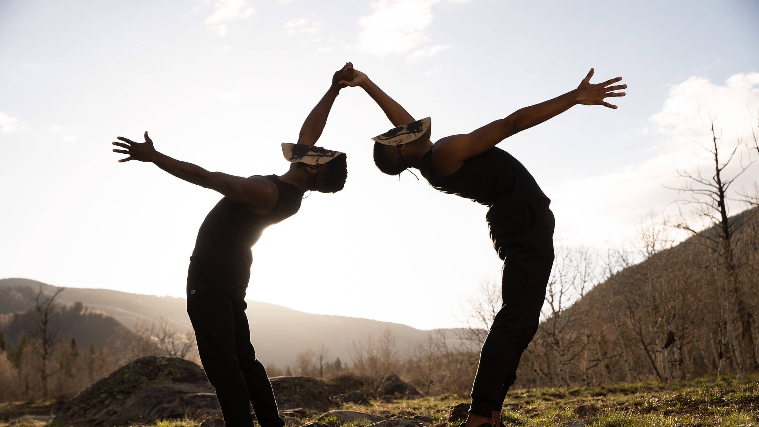 Two men wearing masks bend over backwards towards each other, two of their hands meeting. They're outside and silhouetted against a rising sun.