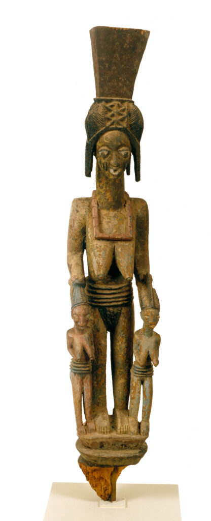 A carved post in the shape of a royal wife standing with hands resting on the heads of her twin daughters.