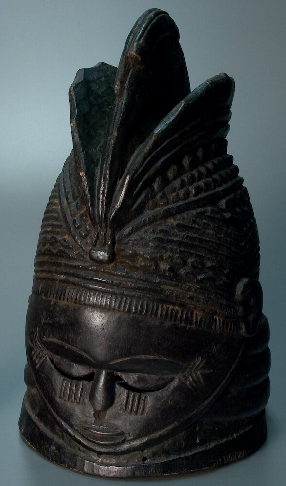A dark wooden mask with a high forehead, downcast eyes, and small mouth.
