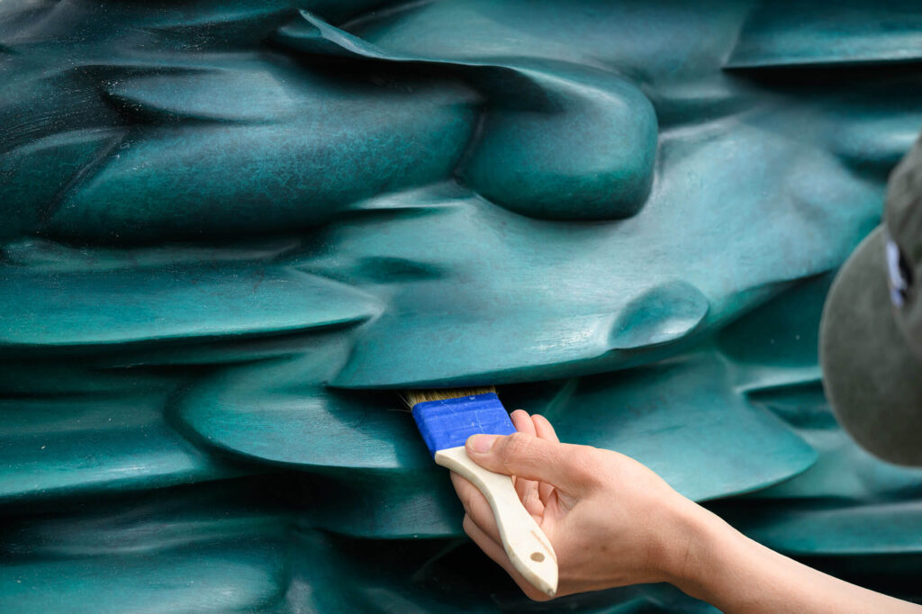 A hand holding a clean paintbrush brushes dirt out from the recesses in a teal sculpture.
