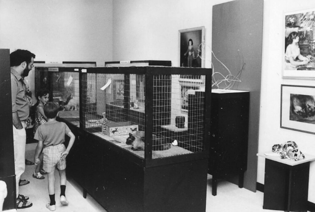 A black and white photo of a parent and two kids exploring a room with art on the walls and several cages with cats in them.