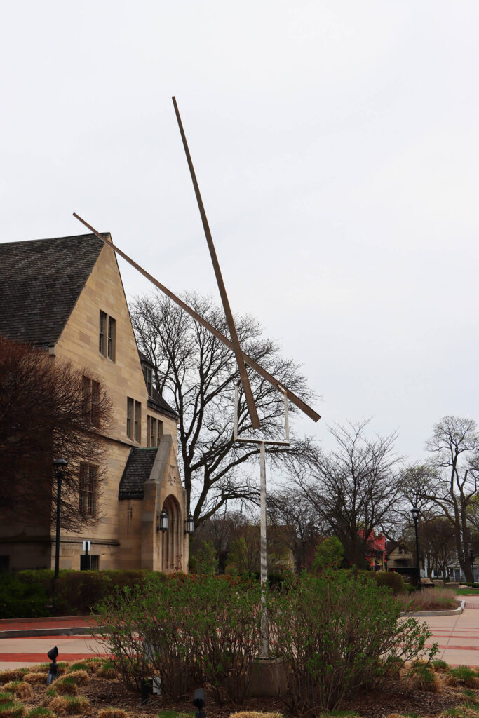 A sculpture with two long, thin poles rotating at the top of another tall, thin pole.