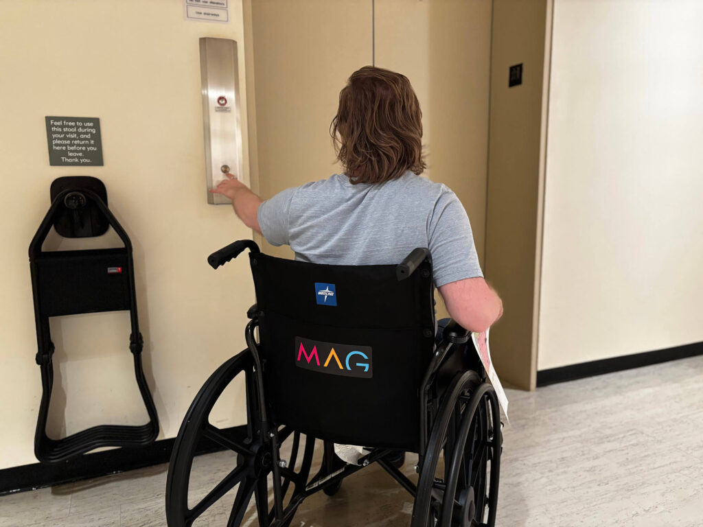 A person in one of MAG's wheelchairs pushing the button for the elevator in the galleries.
