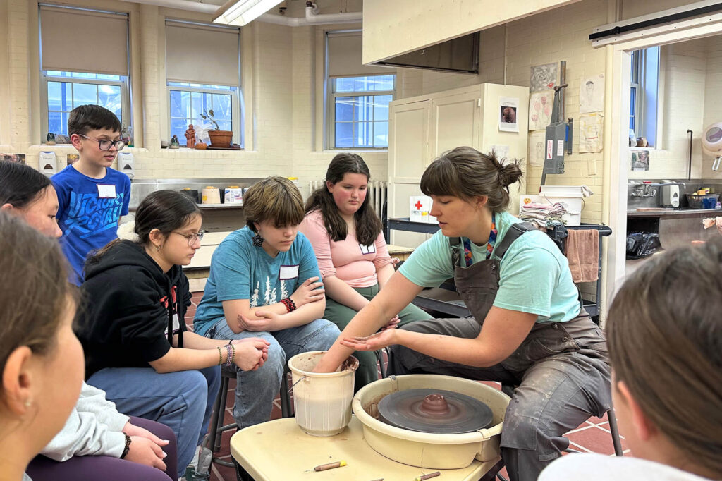 A Creative Workshop instructor gives a pottery wheel demo to a group of campers.
