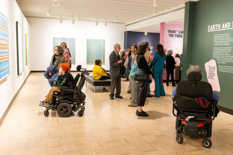 Two people in wheelchairs mingle with other guests and look at art in the galleries at MAG.
