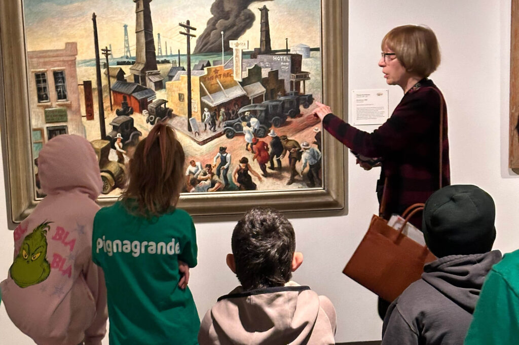 A woman points at a part of a busy painting and talks to four children.