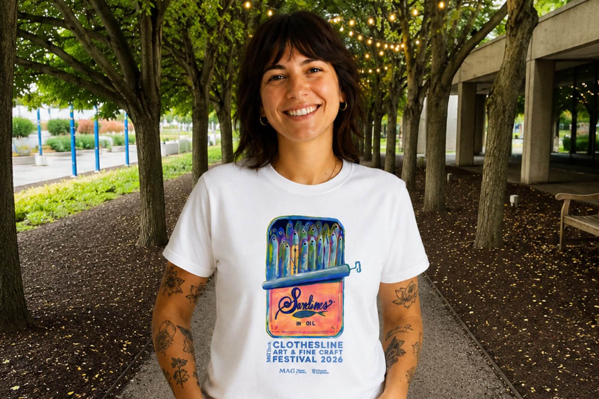 A woman smiling and wearing the 2026 Clothesline tee, which features colorful artwork of a tin of sardines.