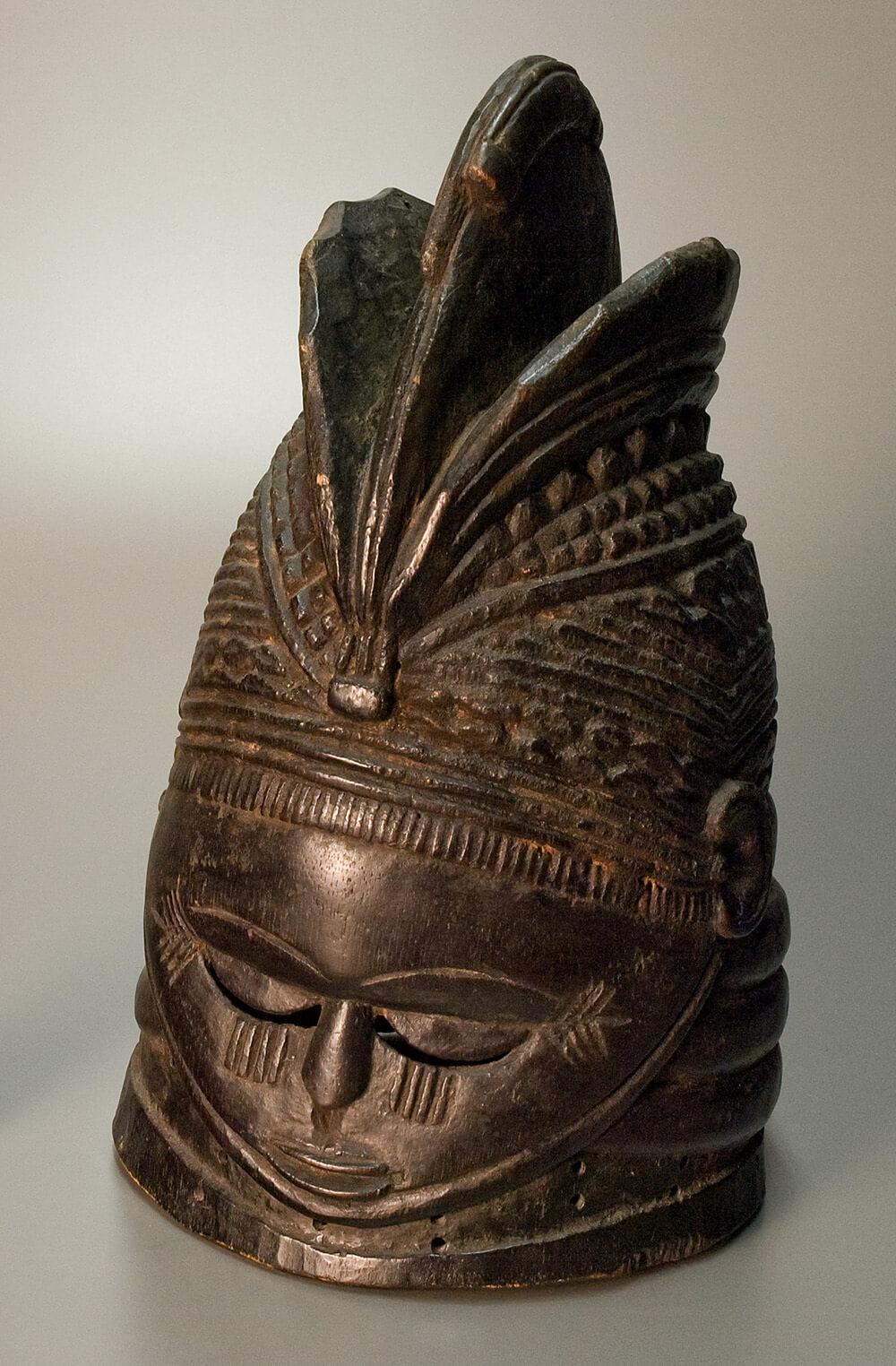 A dark wooden mask with a high forehead, downcast eyes, and small mouth.