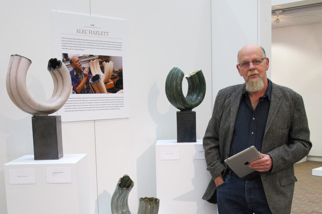 Alec Hazlett standing next to three pieces of his ceramics work in a white gallery.