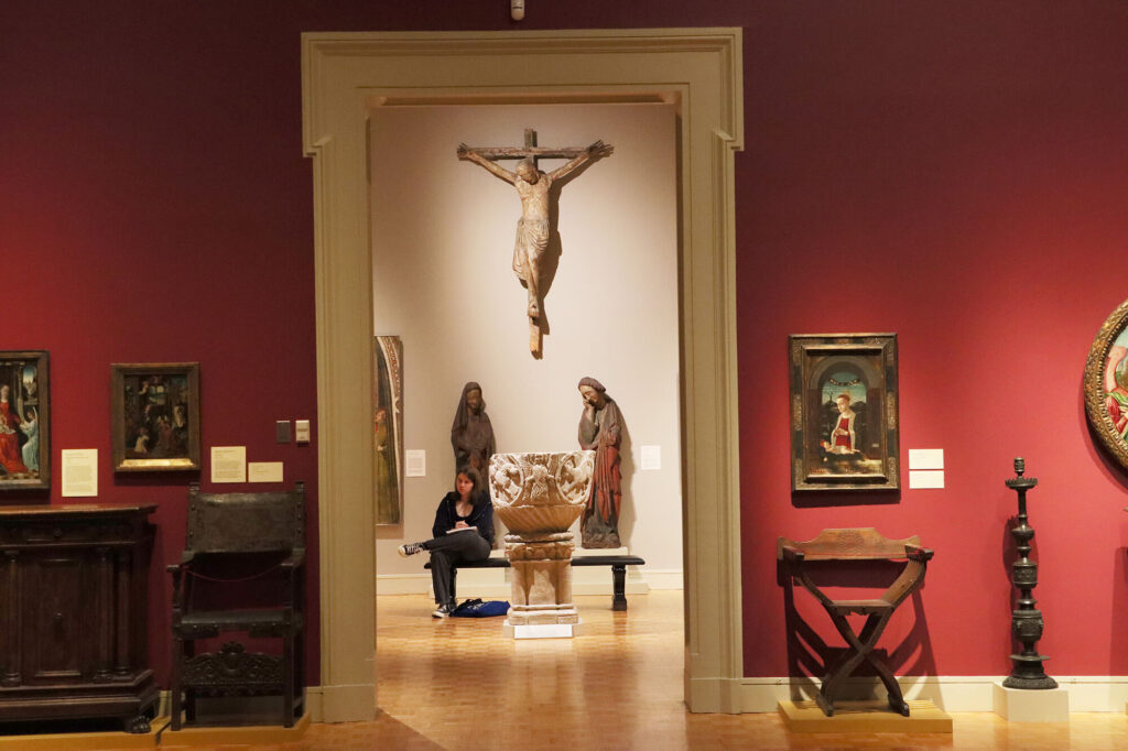 A person sitting on a bench in MAG's Medieval gallery, framed through the door from the Renaissance Gallery.